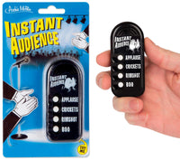 A hand holds the "INSTANT AUDIENCE - Pocket Comedy Sound Machine Noise Maker Toy" by Archie McPhee, a device featuring buttons for sound effects like applause, crickets, rimshot, and boo. The packaging is displayed on the left.