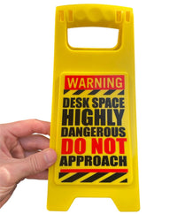 A hand holds the ⚠️ Funny Warning HIGHLY DANGEROUS Desk Sign—an office humor gag joke gift that reads: "WARNING DESK SPACE HIGHLY DANGEROUS DO NOT APPROACH." Perfect for laughs at work!.