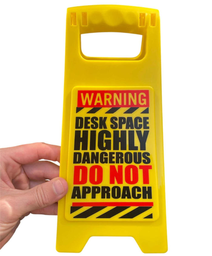 A hand holds the ⚠️ Funny Warning HIGHLY DANGEROUS Desk Sign—an office humor gag joke gift that reads: "WARNING DESK SPACE HIGHLY DANGEROUS DO NOT APPROACH." Perfect for laughs at work!.