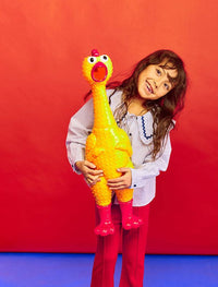 A young girl with long brown hair smiles and holds the GIANT 27" Rubber Chicken—the biggest and loudest squeaky toy—against a bright red background, standing on a blue floor.