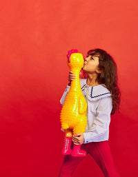 A young girl in a blouse and red pants kisses the 🐔 GIANT 27" Rubber Chicken—the biggest, loudest squeaky toy and a hilarious gag gift—against a red background.