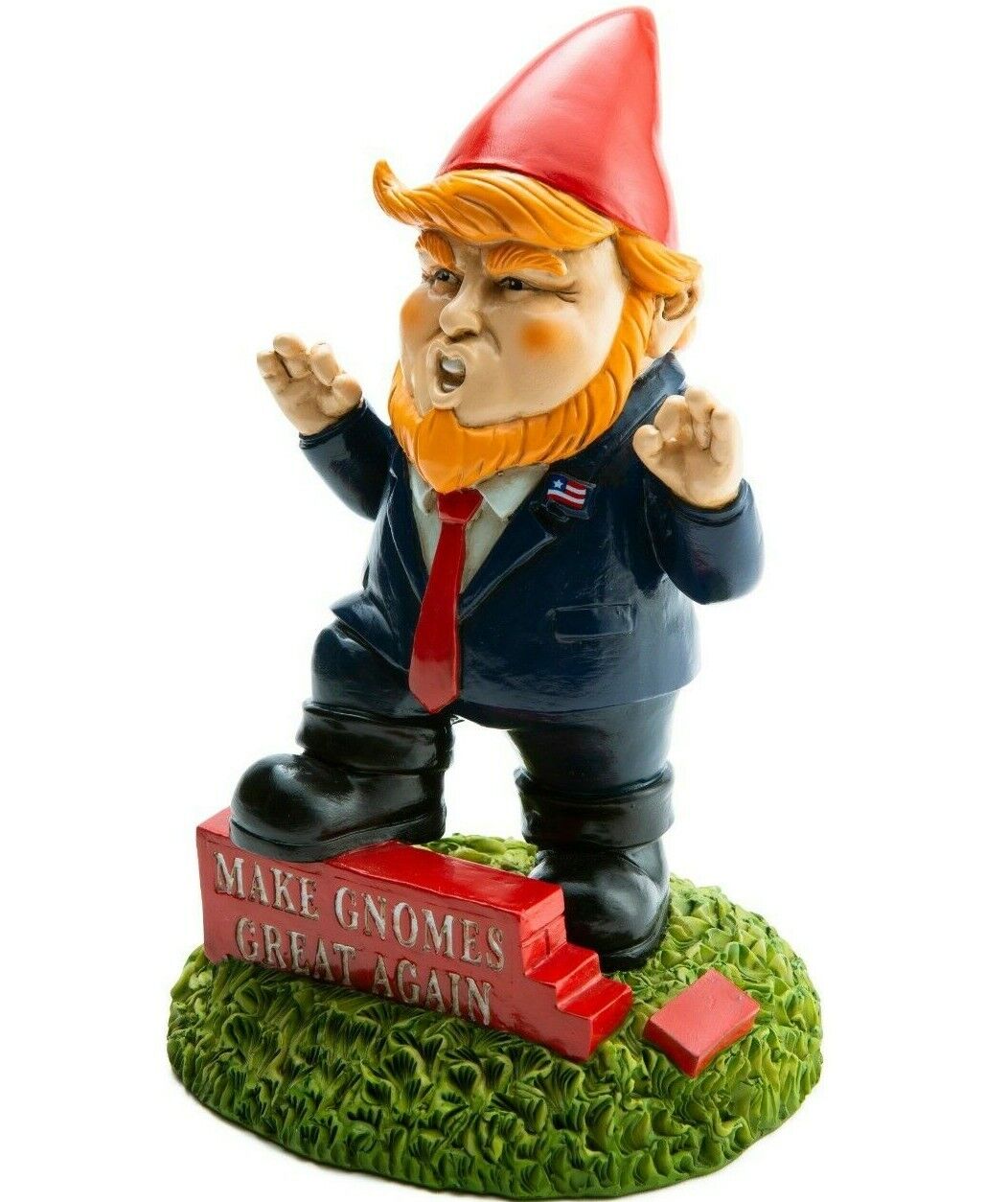 A funny garden gnome dressed in a suit, red tie, and red hat stands on grass next to a small podium with the words "Make Gnomes Great Again." This hilarious Donald Trump garden gnome is sure to be a conversation starter, brought to you by BigMouth Toys.