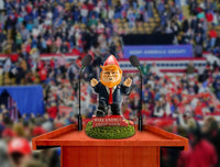 A hilarious DONALD TRUMP GARDEN GNOME from BigMouth, dressed as a political figure resembling Donald Trump, stands behind a podium with two microphones in front of a blurred crowd. The sign on the podium reads, "Make Gnomes Great Again.