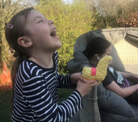 A girl in a striped shirt laughs while holding the SPITTIN' LLAMA Water Pistol Squirt Gun, a funny child spray soaker toy. Next to her sits another person with earphones in, looking down. They are outdoors with greenery in the background.