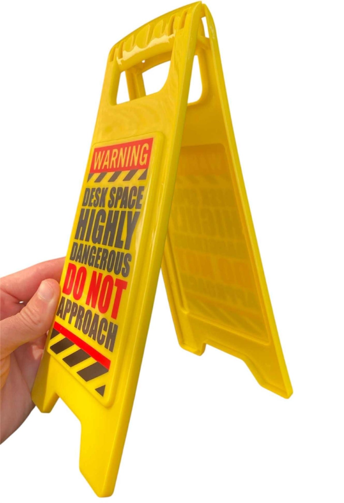 A hand holds the ⚠️ Funny Warning HIGHLY DANGEROUS Desk Sign, which reads "Warning desk space highly dangerous do not approach"—a great office humor gag gift for your workspace.