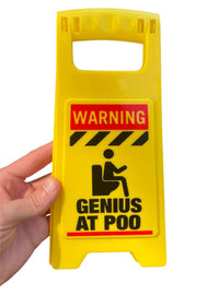 A hand holds the ⚠️ Funny Warning Genius Poo Desk Sign 💩 🚽, featuring "Warning: Genius at Poo" with a toilet illustration—ideal as an office humor gag or potty joke gift.