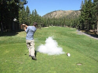 A golfer swings a club on a green course, causing a cloud of dust or smoke to rise from the ground, likely from one of The Ultimate Golf Prank Set's trick golf balls. Trees and mountains stand majestically in the background under a clear blue sky.