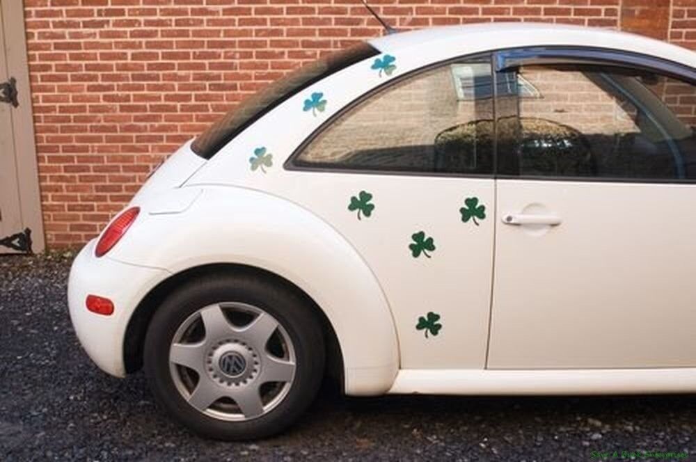 A white Volkswagen Beetle adorned with green four-leaf clover stickers and 12 Shamrock Irish Clover Leaf Car Fridge Kitchen Magnets - Saint Patricks Day is parked next to a brick wall.