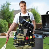 A man in a white shirt wears THE GRILL SERGEANT BBQ APRON, camouflaged with various condiments and utensils, while standing beside a barbecue grill.