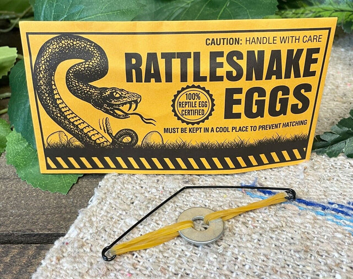 Notice reading "144 Prank Rattlesnake Egg Envelope" with an illustration of a rattlesnake. Includes cautionary text "Handle with care" and "Must be kept in a cool place to prevent hatching." Yellow device shown below. Perfect for adding some excitement to your next party as a rattlesnake eggs prank!