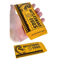 A hand holding a yellow envelope labeled "Rattlesnake Eggs" with a caution notice, perfect for the classic rattlesnake eggs prank. Another identical envelope lies underneath, making it an ideal choice for party favors or buying in bulk from the 144 Prank Rattlesnake Egg Envelope - Novelty Gag Joke Gift Toy Wholesale Lot.