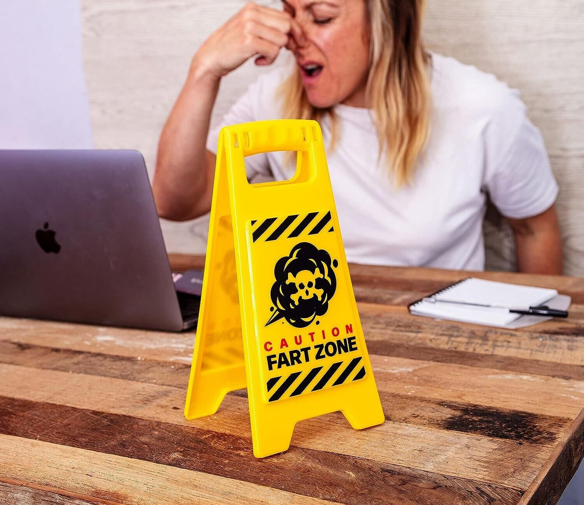 A woman pinches her nose in discomfort at a table with a laptop, while the "Fart Zone Desk Warning Sign Hilarious Office Gift Accessory - Caution Sign GaG" sits in front of her—clearly, someone's toilet humor has turned into an unforgettable gag gift.