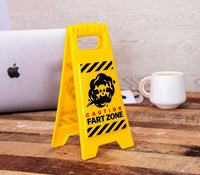 On a wooden table sits the "Fart Zone Desk Warning Sign Hilarious Office Gift Accessory - Caution Sign GaG," sporting a yellow caution sign that reads "Caution Fart Zone" with a graphic of a cloud emitting gas, making it an ideal gag gift. Surrounding it are a laptop, a white mug, and other desk items.