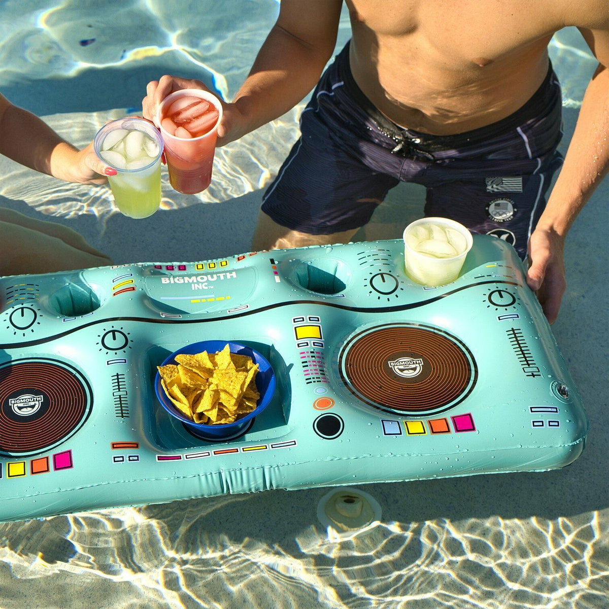 Two people in swimwear enjoy drinks by the pool, placing them on a DJ Table Beverage Float from BigMouth that also holds a bowl of chips, turning it into the perfect portable party accessory.