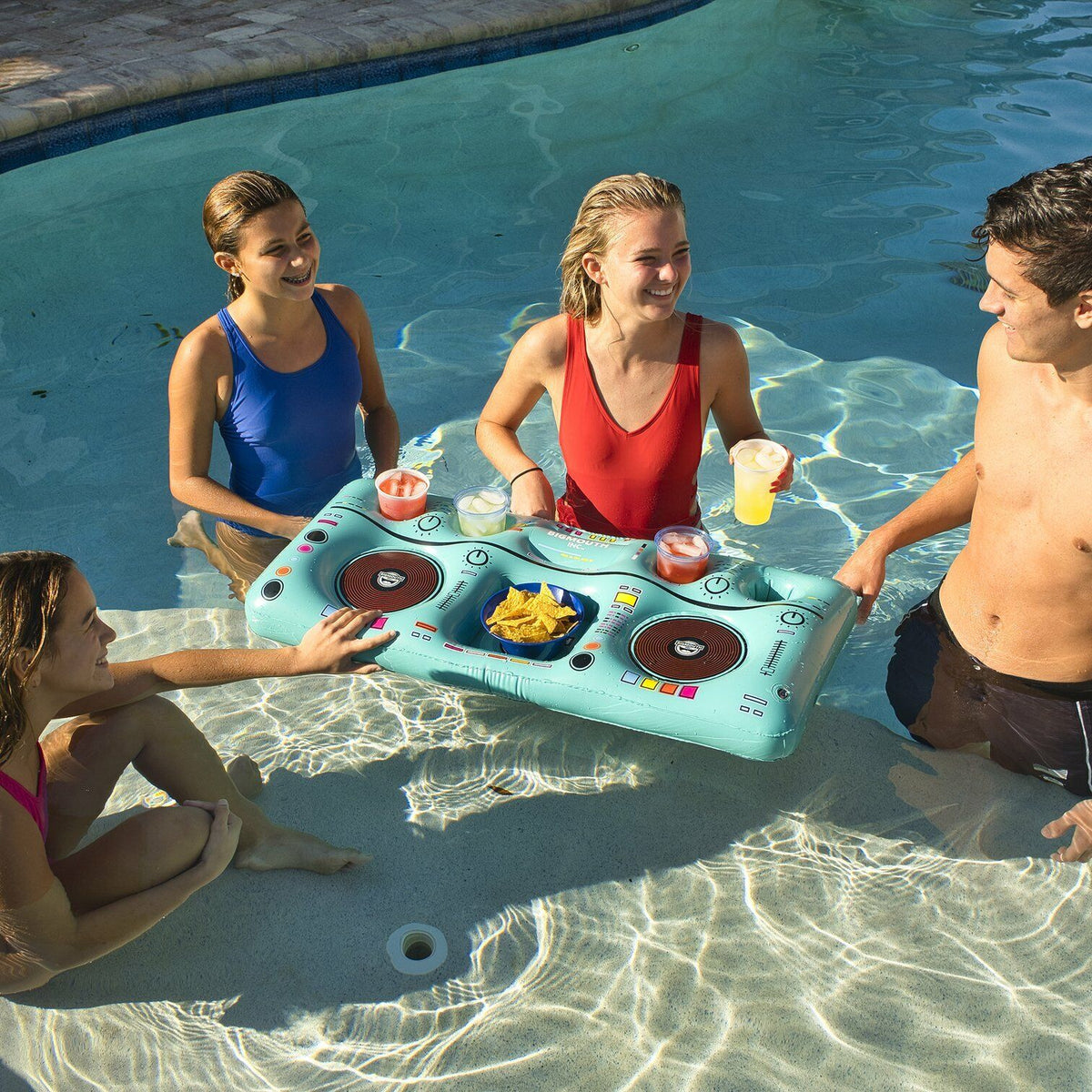 Four people in swimsuits are in a swimming pool, gathered around the DJ Table Beverage Float - Party Buffet Bar Beer Food Pool Cooler by BigMouth. This inflatable float, designed to resemble a DJ turntable, holds drinks and snacks, making it the perfect portable party accessory.