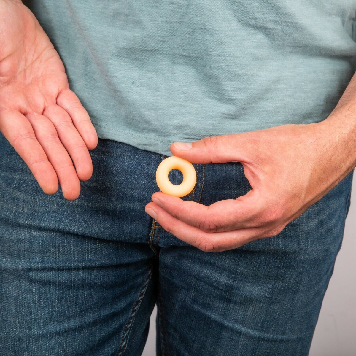 A person in a blue shirt and jeans is holding a ring-shaped object near their waist with one hand, while their other hand is open, palm facing up. They're likely demonstrating The MINI MICRO ITTY-BITTY Willy Weener Soap—a humorous yet practical gag gift aimed at improving personal hygiene from BigMouth.