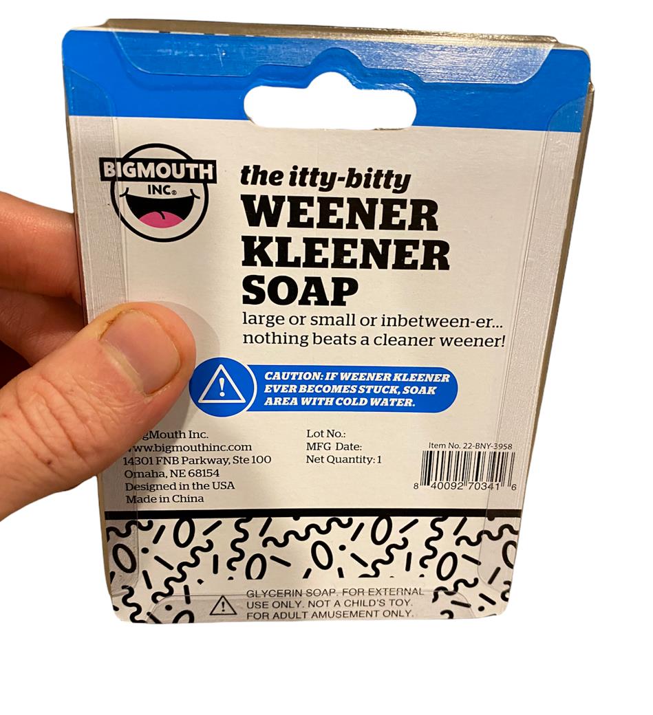 A hand holding "The MINI MICRO ITTY-BITTY Willy Weener Soap" packaged product by BigMouth Inc. The packaging features a cartoon and cautionary text about the gag gift, adding a playful touch to personal hygiene.