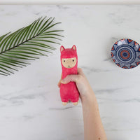 A hand holds the CALMA LLAMA Squishy Fidget Durable Squish in pink on a marble surface. Nearby, a green palm leaf and a small decorative bowl complete this tranquil scene, making it a perfect anti-anxiety gift.