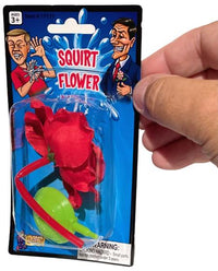 A hand holding a packaged novelty toy labeled "Classic Lapel SQUIRT FLOWER Clown Rose Joke Prank Squirting Water Formal Costume," featuring a red flower with a bulb. The packaging shows a cartoon man squirting water from the flower at another person—a classic clown gag perfect for any prank gag gift occasion.