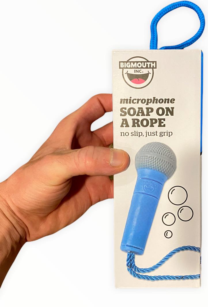 A hand holds a boxed "Microphone Soap On Rope - Rock on while cleaning up your butt!" by BigMouth Inc. Perfect for shower concerts, the packaging shows a blue microphone-shaped soap with a rope attached and text that reads "no slip, just grip.