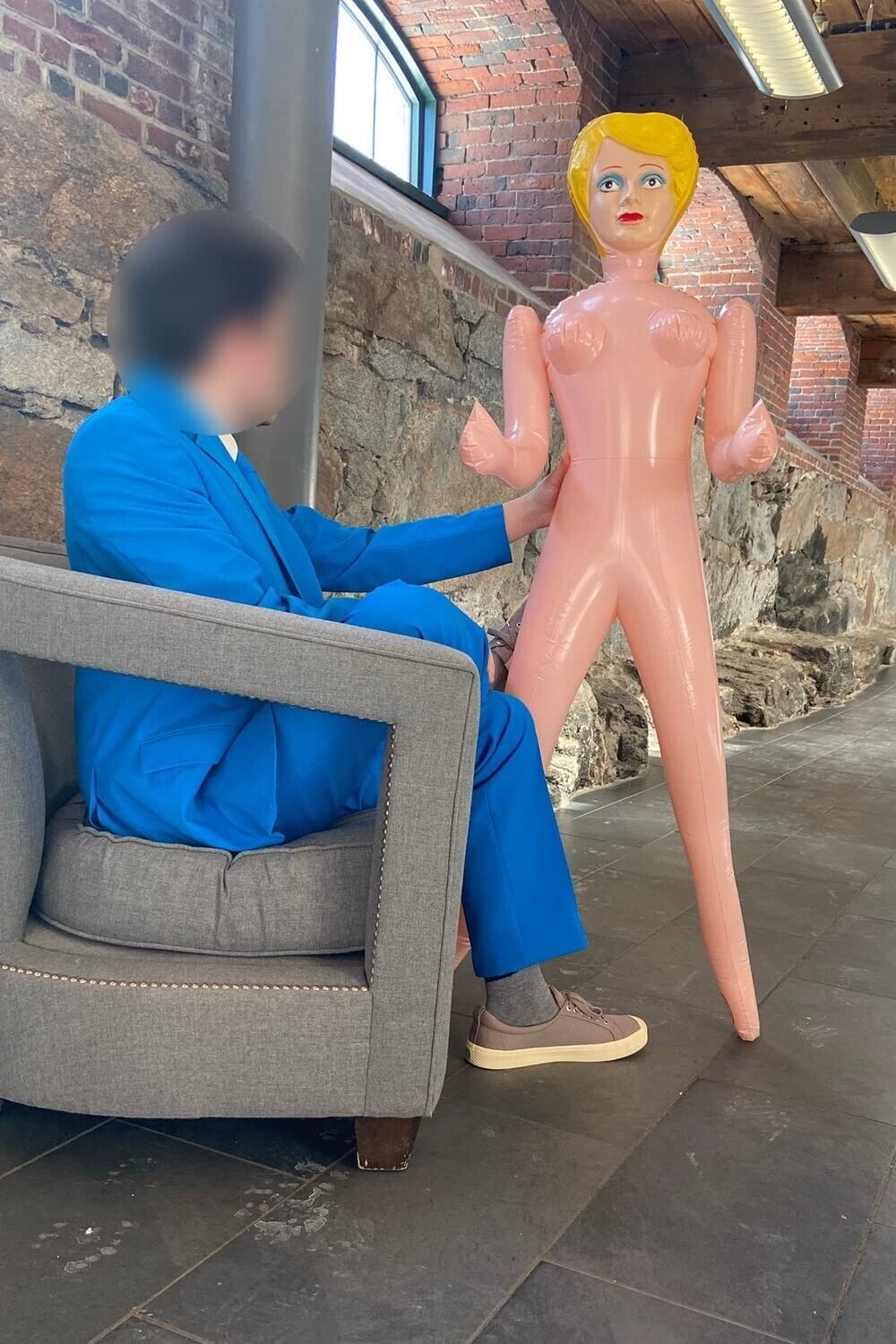 A person wearing a blue suit sits in a chair next to Inflatable Judy from the "Inflatable Judy + John Inflate a Date Bachelor/Bachelorette Party Blow Up Dolls" collection, creating an intriguing scene in the brick interior setting.