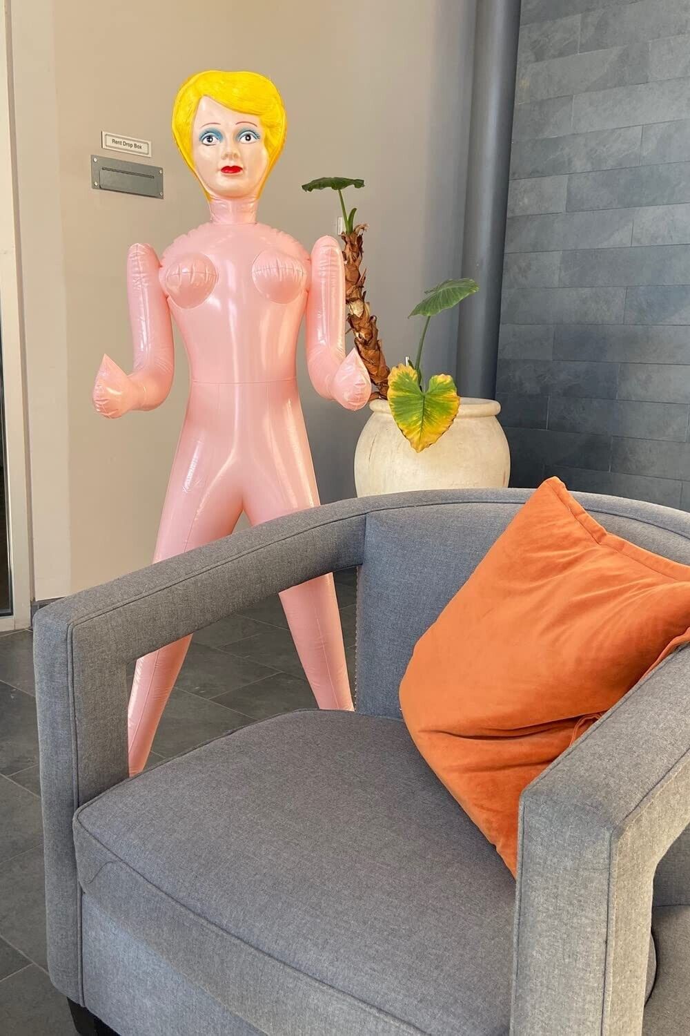 A life-size Inflatable Judy from the Inflate a Date Bachelor/Bachelorette Party Blow Up Dolls collection, featuring yellow hair, stands behind a gray armchair with an orange cushion, adjacent to a potted plant.