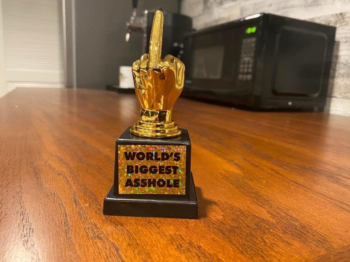 A funny World's Biggest AS%HO#E Trophy with a golden hand showing the middle finger stands proudly on a wooden table. The base of this humorous gift reads "WORLD'S BIGGEST AS%HO#E" in colorful letters—a perfect gag gift for someone with a sense of humor.