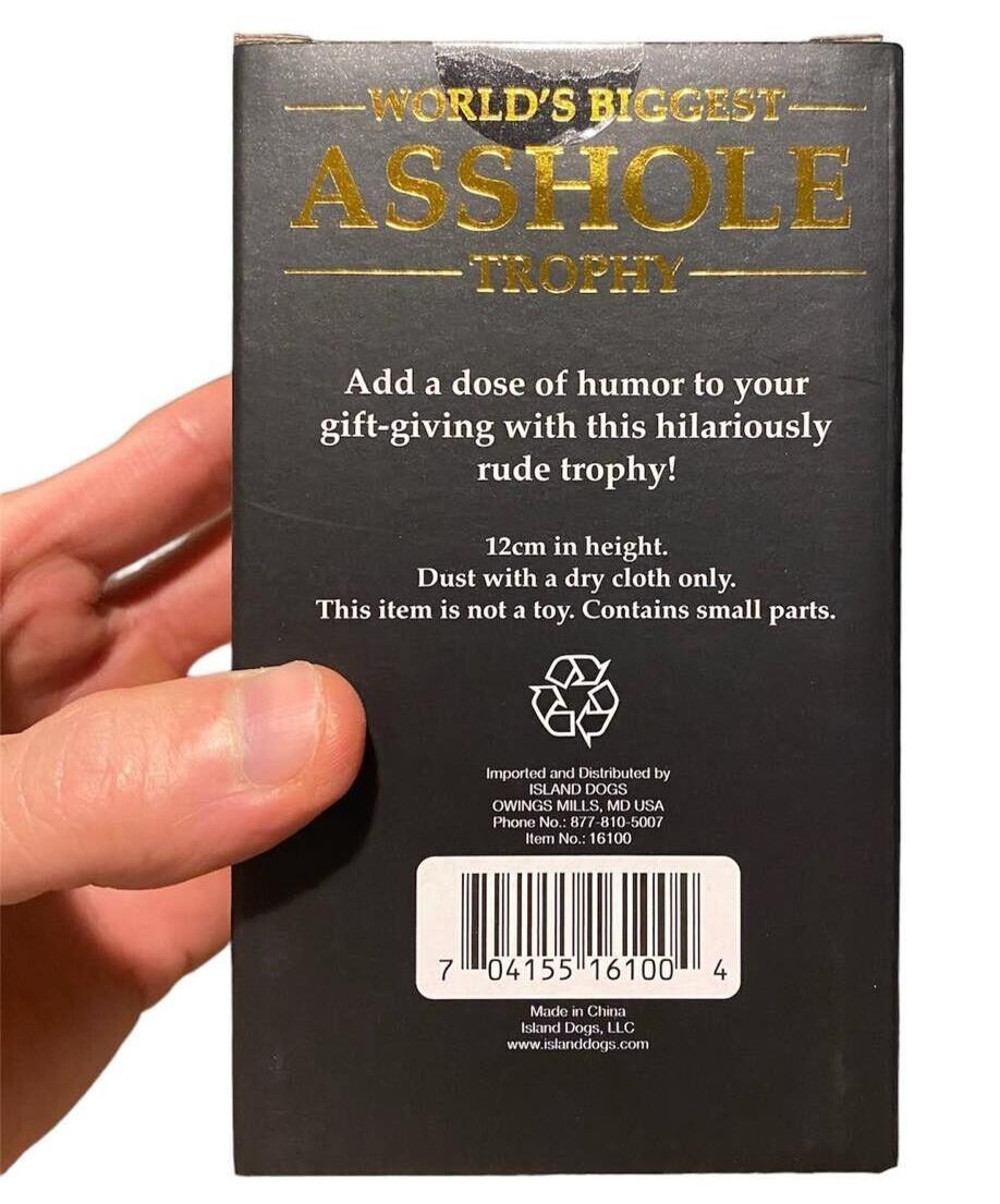 Close-up of packaging for the "World's Biggest AS%HO#E Trophy Middle Finger F-U- Golden Award," a funny trophy held by a hand. Text highlights its humor, height (12 cm), and a warning about small parts. Barcode and distributor information visible, making it the perfect gag gift.