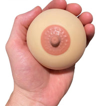 A hand holds a round object resembling a breast with a nipple, ideal as a gag gift or for honing your juggling skills. The 2pk Stress Boobs - Squeeze Boobies Feels Real! Adult Novelty Breast Man Toy Gift is not just entertaining but also great for relieving tension and highlighting your playful nature.