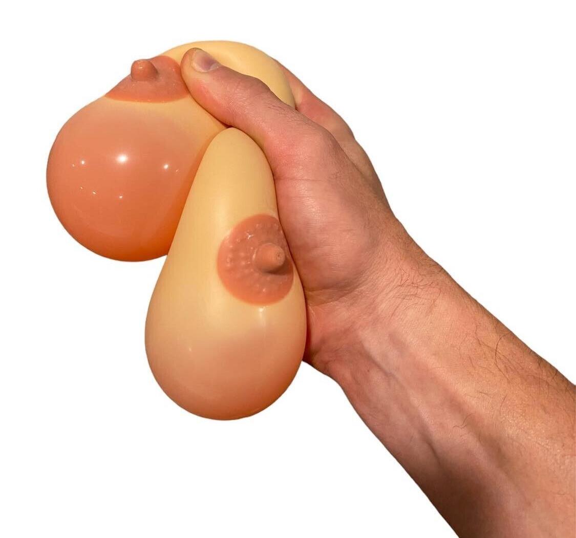 On a white background, a hand squeezes two silicone stress balls cleverly named "2pk Stress Boobs - Squeeze Boobies Feels Real! Adult Novelty Breast Man Toy Gift," complete with visible nipples. They are perfect for refining your juggling skills or offering as an amusing gag gift.