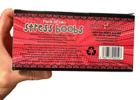 A hand holds a rectangular package labeled "2pk Stress Boobs - Squeeze Boobies Feels Real! Adult Novelty Breast Man Toy Gift," making it an ideal gag gift. The packaging, adorned with playful warning texts and branding details, stands out against the red background.