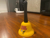 A yellow "FART ALERT" Hand Bell, complete with a black handle, rests on a wooden surface. This quirky bell, adorned with the text "FART ALERT" and a humorous illustration of a person farting, serves as an entertaining gift or a playful warning system.