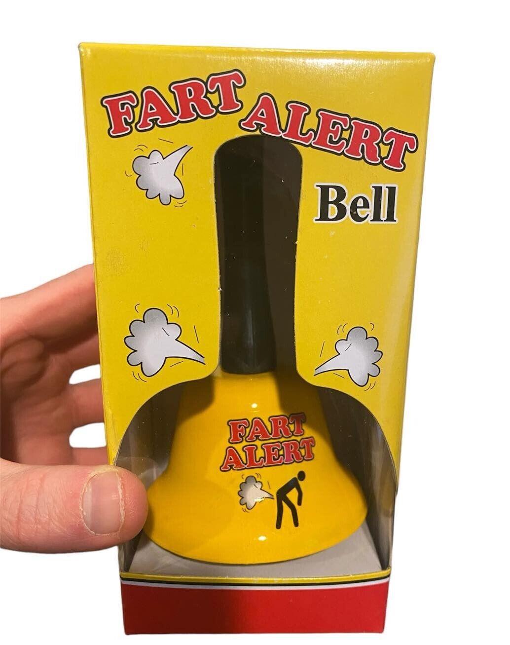 The yellow "The FART ALERT Hand Bell" in packaging adorned with illustrations of fart clouds and a stick figure expelling gas. A hand is holding the box, highlighting this hilarious warning system—making it an ideal gift for pranksters everywhere.