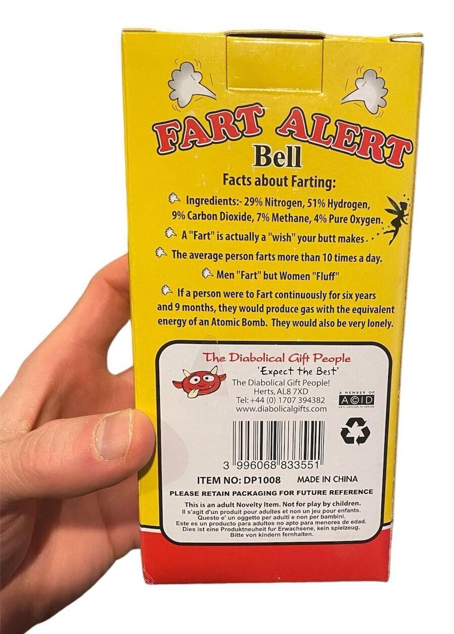 A person holding a yellow box with humorous text about farting and a barcode at the bottom. The product, labeled "The FART ALERT Hand Bell - Funny Joke GaG Gift - Farting Stink Bomb Warning" from "The Diabolical Gift People," is an excellent gift and serves as the perfect warning system.