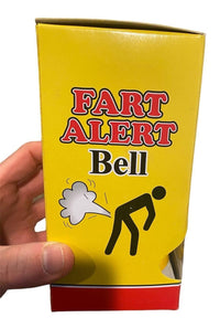 A yellow box labeled "The FART ALERT Hand Bell - Funny Joke GaG Gift - Farting Stink Bomb Warning" features a humorous illustration of a person bending over with a cloud coming from their rear end. A hand is holding the Fart Alert hand bell on the left side, making it a great gift and an amusing warning system for any occasion.