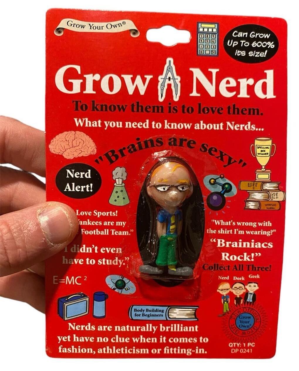 A hand holds a "SET OF 3 Grow your own DORK - NERD - GEEK" toy package that features a nerd figurine along with humorous geek-related text and images. The packaging claims the toy can grow up to 600% its size in water.