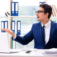 A man in a blue suit and glasses sits at a desk, expertly tossing three black Magnetic Rattlesnake Eggs - Magic Fidget Stress Toys in the air. He is surrounded by an office setting, with shelves of neatly arranged binders behind him.