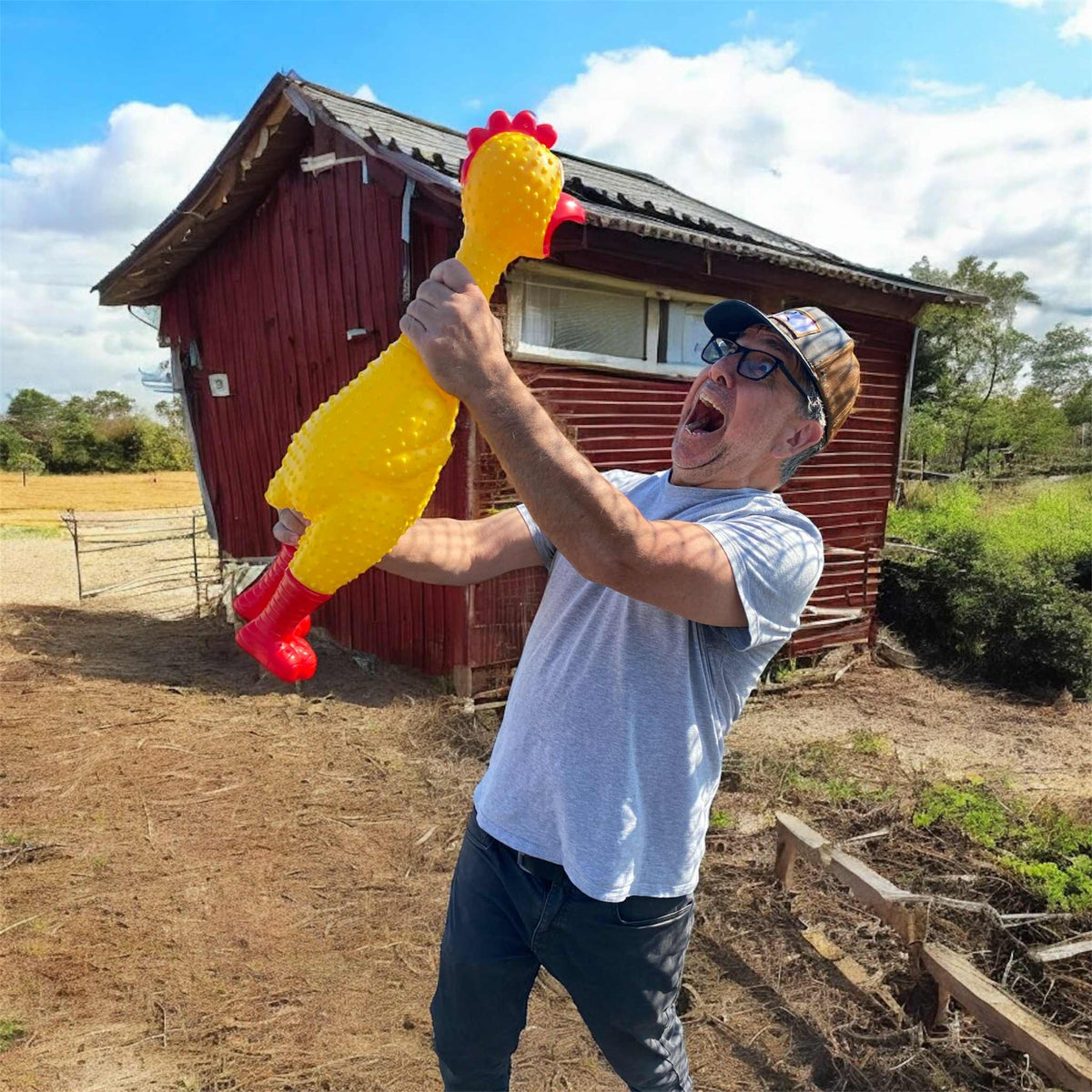 A man in glasses and a cap stands outside by a red wooden building, holding the 🐔 GIANT 27" Rubber Chicken—the biggest and loudest squeaky toy—and looks at it with an open-mouthed expression.