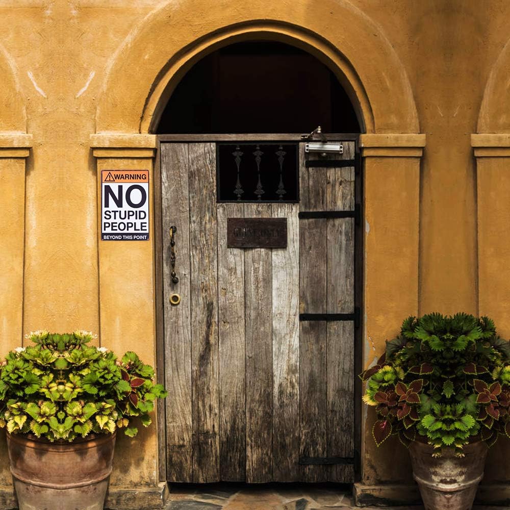 Highlighting novelty decor at its best, this metal tin sign reading "Warning: NO STUPID PEOPLE Beyond This Point" adds a touch of humor to any space. Positioned prominently on a wooden door between two potted plants against a vibrant yellow wall, it is sure to evoke smiles and set the tone for what lies ahead.