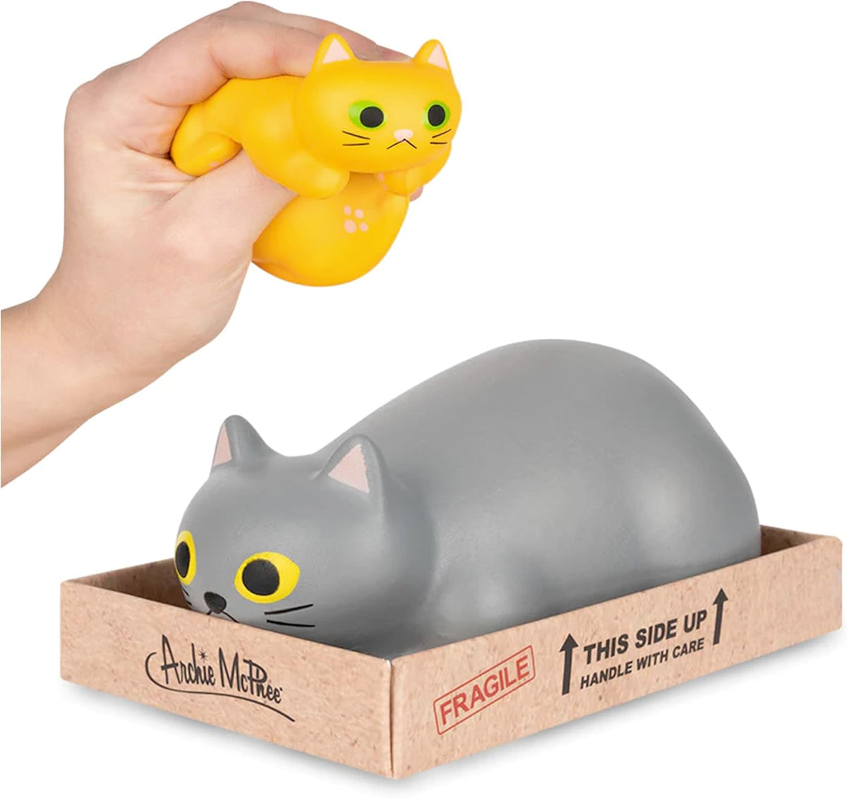 A hand squeezes a squishy yellow stress toy from the Purrrly Kitty Cat in a Box collection by Archie McPhee above a larger gray cat toy in a box labeled "Fragile" and "This side up.