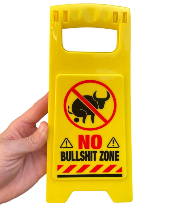 A hand holds the ⚠️ Funny Warning No Bullshit Zone Desk Sign with a crossed-out bull graphic and bold red and black text—an office humor gag joke and funny gift.