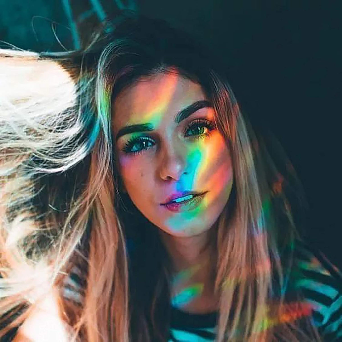 A woman with long hair is enveloped in the captivating rainbow hues cast by the PRISM - Fun Educational Science Rainbow Light of Colors by Archie McPhee, highlighting her face and striped shirt.