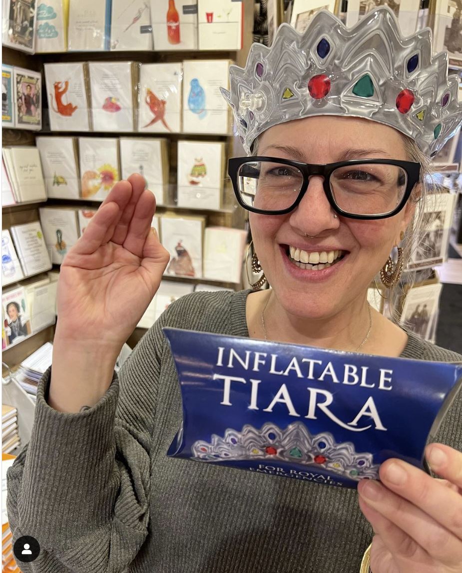 Amidst royal emergencies, an individual with glasses and an Emergency Inflatable Tiara - Royal Crown Princess Queen Headband by Archie McPhee confidently holds its packaging while standing in a store filled with greeting cards. This whimsical gag gift is sure to add a touch of humor to any occasion.