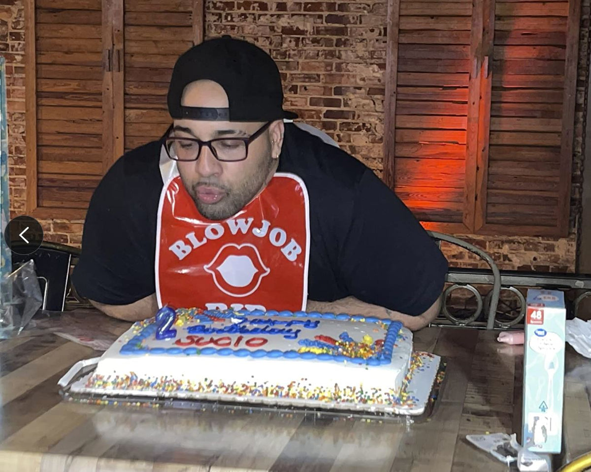 A man in a backwards cap, wearing the Blowjob Bib Bachelorette Party Favor 💋 adult gag gift, blows out candles on a decorated birthday cake at an indoor wooden table.