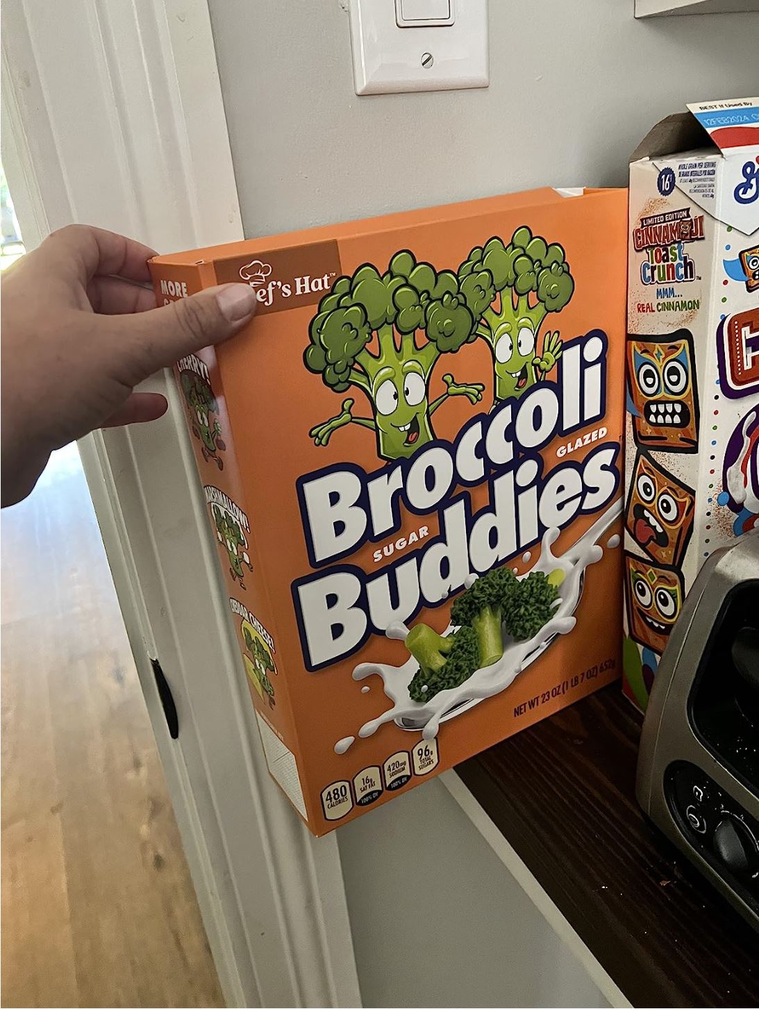 A hand holds the 🥦 Prank Cereal Box – Broccoli Buddies Gag Gift Box | Funny Joke Prank Present🎁 with cartoon broccoli characters, perfect for a prank or gag gift, displayed next to other cereal boxes on a kitchen counter.