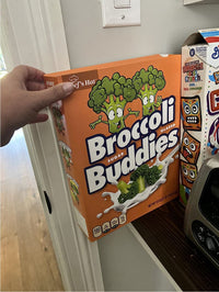 A hand holds the 🥦 Prank Cereal Box – Broccoli Buddies Gag Gift Box | Funny Joke Prank Present🎁 with cartoon broccoli characters, perfect for a prank or gag gift, displayed next to other cereal boxes on a kitchen counter.
