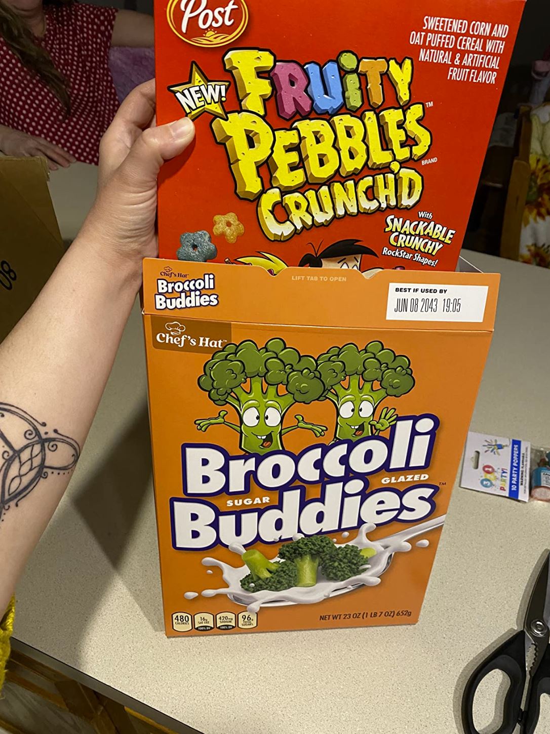 A hand holds two cereal boxes on a countertop—a Fruity Pebbles Crunch'd prank box stacked on top of a 🥦 Prank Cereal Box – Broccoli Buddies Gag Gift Box | Funny Joke Prank Present🎁.