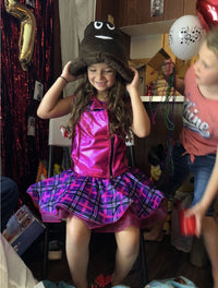 A young girl in a pink and purple dress sits on a chair putting on the 💩 Poop Emoji Hat – Funny Gag Gift & Hilarious Party Costume Accessory, getting ready for the party, while another child stands nearby holding beaded necklaces.