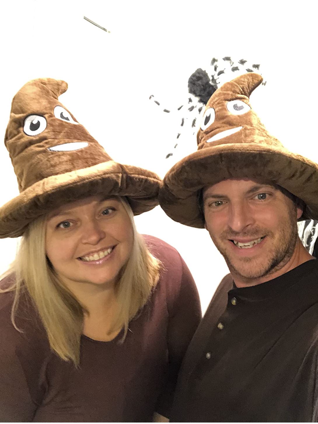Two adults smile, each wearing the 💩 Poop Emoji Hat – Funny Gag Gift & Hilarious Party Costume Accessory, a brown plush hat with eyes and a grin—perfect for adding laughter to your next costume party.