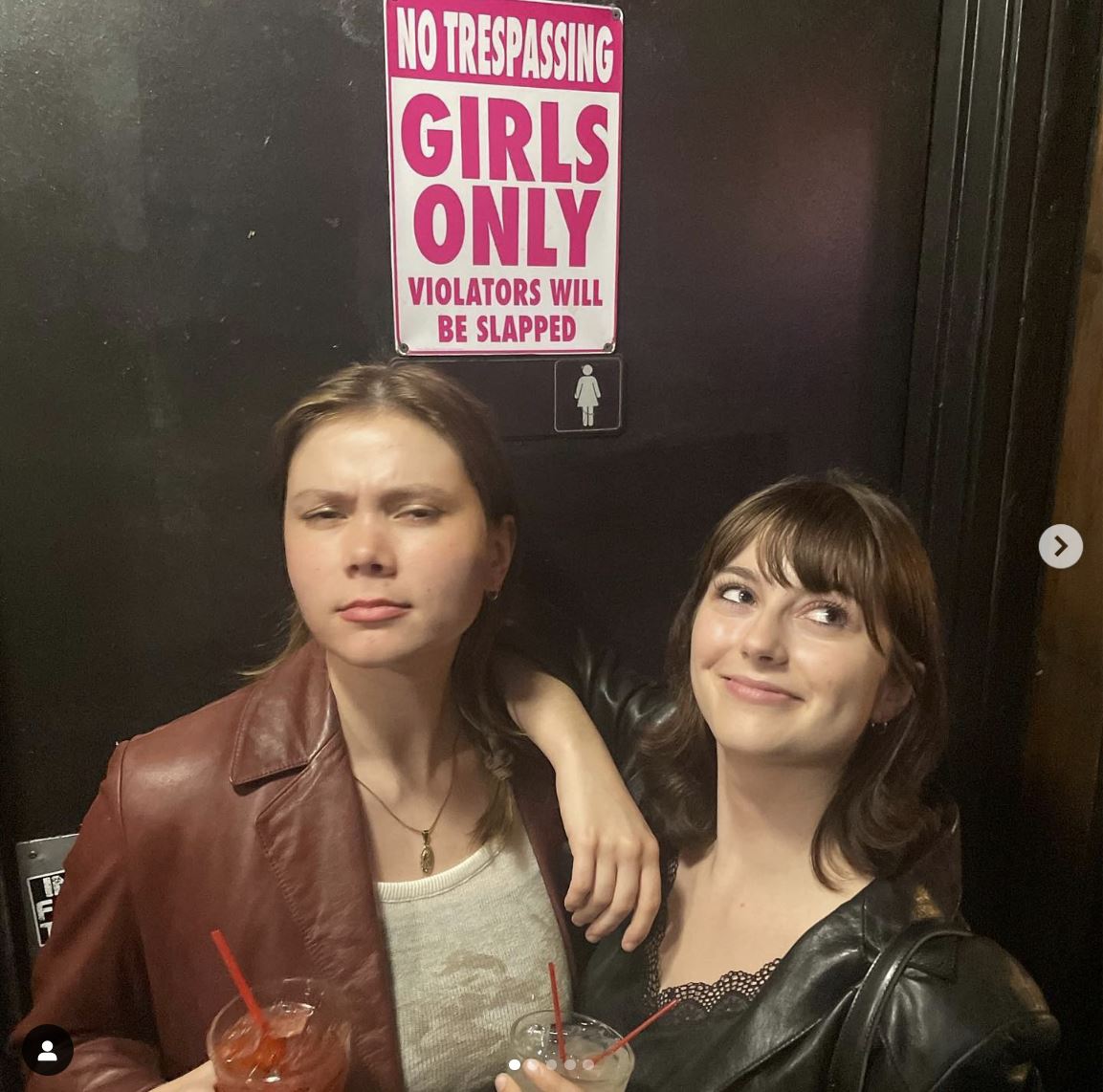 Two people holding drinks stand under a "Girls Only" door sign, enhanced by a humorous wall decor featuring the metal tin sign "No Trespassing: Girls Only Violators Will Be Slapped." One person is serious while the other smiles, contributing to the scene's light-hearted atmosphere.